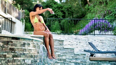 Skinny young ebony babe in tiny yellow string bikini sits poolside dangling bare feet in water