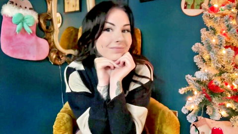 Tattooed emo girl in festive sweater smiles cutely with hands on chin in Christmas room
