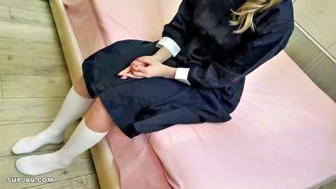 Blonde schoolgirl in black sailor uniform sitting on pink bed with legs apart and hands clasped teasing