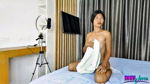 Topless Asian babe with perky tits sits on bed holding towel over pussy teasing