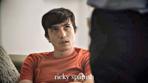 Young man in orange shirt sitting on couch, Ricky Spanish text overlay