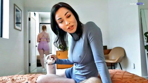 Asian beauty in sheer blue bodysuit with visible hard nipples sits on bed eyeing camera as blonde walks away showing ass
