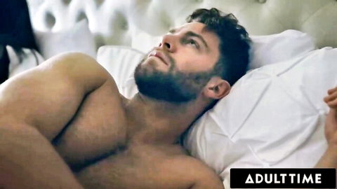 Muscular bearded man lying shirtless on bed in steamy adult scene featuring Angela White and cast