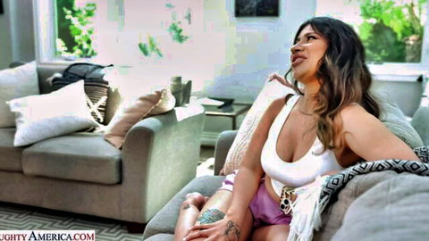 Curvy brunette MILF with tattoos lounging seductively on couch in tight white tank top showing massive cleavage and pink shorts