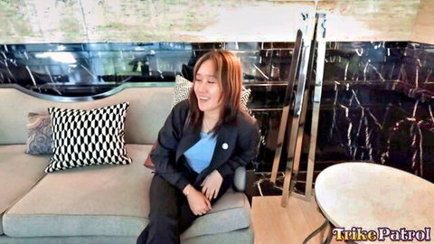 Smiling Asian babe in blazer and blue top sits on couch teasing with naughty grin