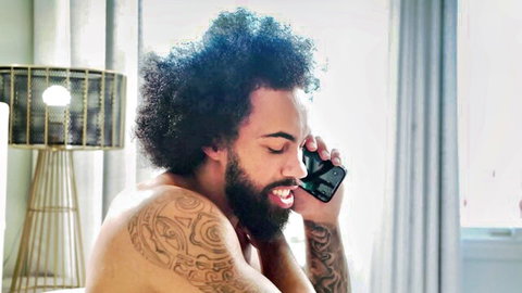 Shirtless tattooed man with blue curly hair smiling on phone call