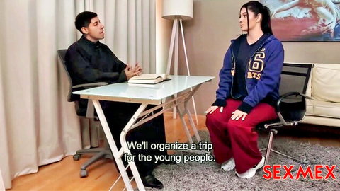Curvy Latina in blue jacket and tight red pants sits talking to man at glass table
