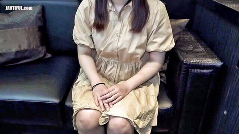 Shy Asian girl in short beige dress sitting on black couch with hands in lap