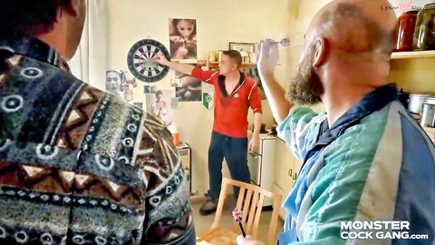 Bald bearded man watches guys play darts in cluttered kitchen on Monster Cock Gang site