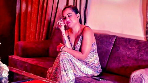 Seductive South Asian woman with arm tattoos teasing deep cleavage in floral dress on couch