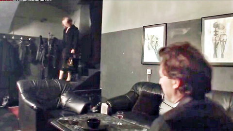 Elegant blonde MILF in tight black dress descends dark stairs toward suited man in leather lounge for femdom tease