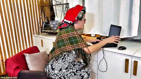 Mature Mel Oliveira at desk in floral dress and plaid shawl