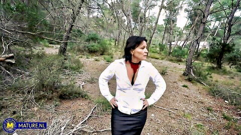Naughty mature MILF with short hair stands hands on hips flashing deep cleavage and red bra in open white blouse and tight black skirt in forest