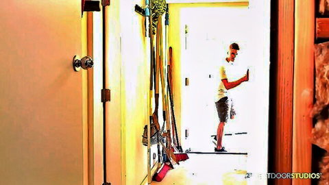 Young athletic Caucasian guy in white tee and shorts stands in sunny doorway amid brooms and mops