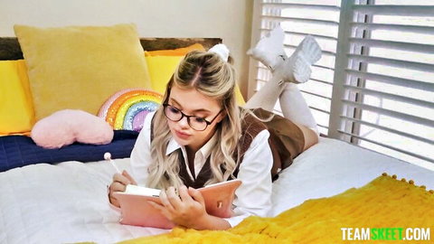 Blonde schoolgirl in glasses and thigh-high socks lies on bed with legs up writing in notebook teasing