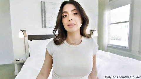 Brunette beauty with big tits smiling seductively in tight white top on bed