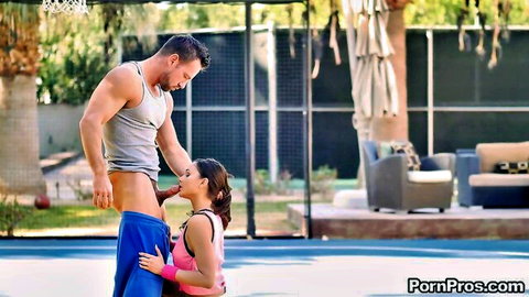 Skinny brunette kneels mouthing tall muscular guy's cock bulge on outdoor basketball court