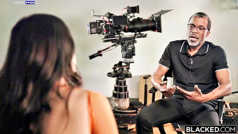 Brunette woman in orange top interviews older black man with glasses on BLACKED professional porn set