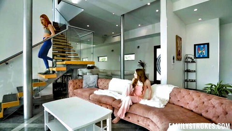 Redhead in tight blue leggings descends glass stairs to seductive brunette lounging on pink couch in luxurious home.
