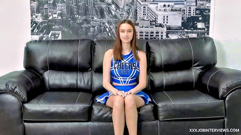 Skinny teen cheerleader in tight blue uniform sits smiling on couch with small tits visible for casting interview