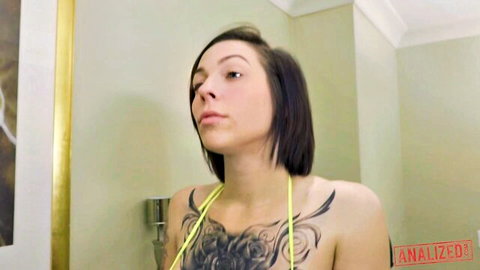 Tattooed brunette with huge tits exposed in yellow harness posing topless in bathroom mirror