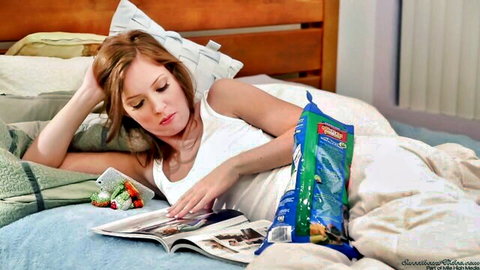 Sexy redhead in tight white tank top lounging in bed reading magazine with chips and strawberries