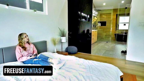Blonde gamer girl in pink sweater and tight jeans sits on bed holding controller smiling playfully
