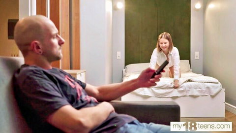 Bald man on couch holds remote watching young blonde teen bending over bed in white blouse POV