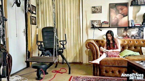 Anna De Ville lounging in red dress amid BDSM setup, seductive pose