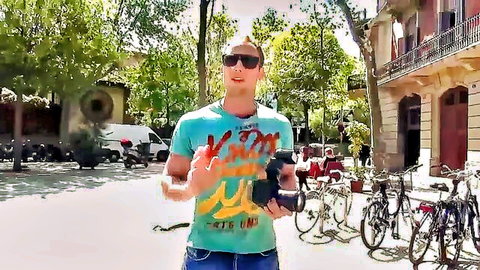 Man in turquoise KMM shirt and sunglasses holding camera gesturing on sunny urban street