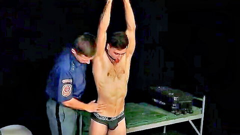 Young cop in uniform gropes restrained tattooed muscular prisoner in underwear