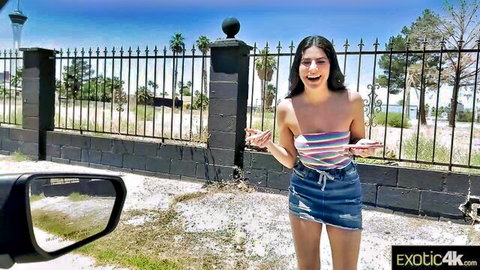 Excited Latina babe in striped tube top and ripped denim shorts smiling outdoors by fence