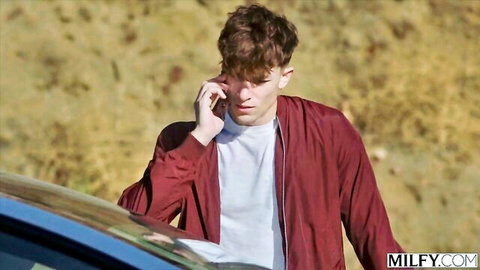 Young curly-haired guy in red jacket talking on phone by blue car on MILFY.com