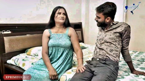 Indian cougar in green dress sits intimately with man on bed, teasing affair vibes