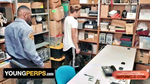 Black cop in uniform stands behind young white twink suspect in cluttered storage room