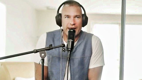 Bald man with headphones speaking into microphone wearing vest and white shirt in podcast setup