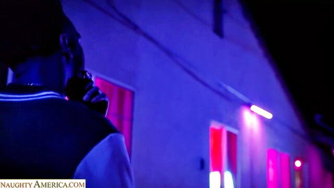 Black man in bomber jacket and hat talking on phone under purple neon lights at night