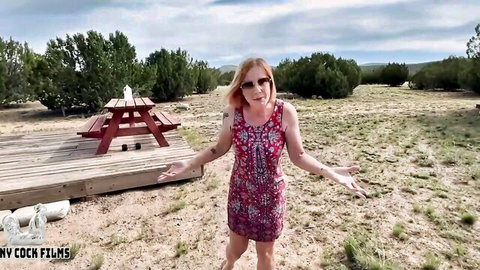 Jane Cane in red patterned dress, arms outstretched outdoors at picnic table