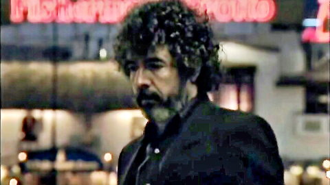 Curly-haired man in dark suit stands outside neon-lit hotel at night