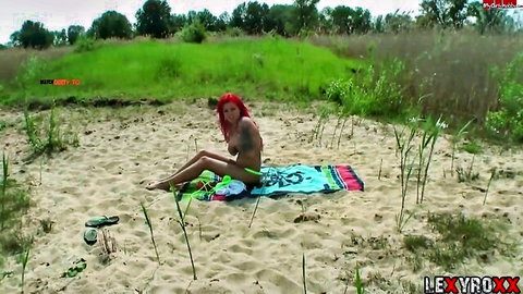 Nude redhead with tattoos sits legs spread on beach towel teasing pussy outdoors