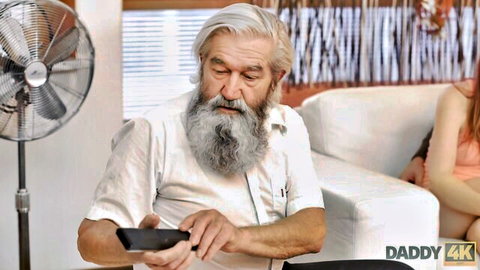 Mature bearded man gazes at phone with young redhead nearby on couch