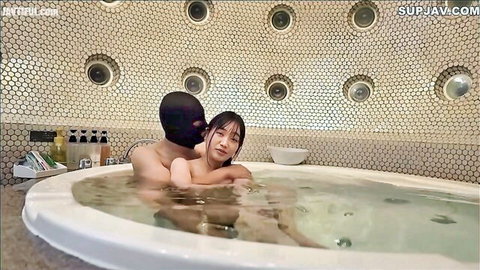 Slim Asian woman embraced by masked black man in steamy hot tub embrace