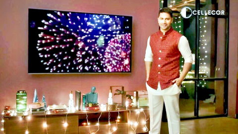 Handsome Indian man in red vest and white outfit posing confidently in festive room with fireworks on TV
