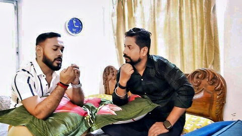 Two bearded Indian men in shirts sit pensively on bed with pillows in bedroom.