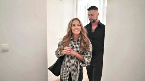 Smiling blonde in checkered blouse holds coffee mug as bearded hunk sneaks up behind her in hotel hallway foreplay tease