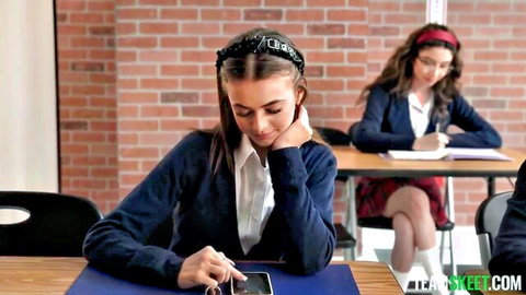 Two sexy schoolgirls in tight uniforms sit at classroom desks, one checking phone while other studies intently