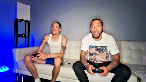 Two muscular black guys excitedly playing video games on white couch in blue-lit room