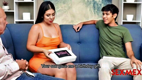 Curvy stepmom in tight orange dress holds birthday gift from son while sitting with husband on living room couch