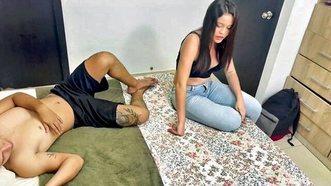 Busty Latina in black bra and jeans sits beside shirtless tattooed guy in low shorts lying on floor mat