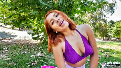 Tanned redhead teen in skimpy purple bikini top smiles seductively over shoulder showing deep cleavage outdoors